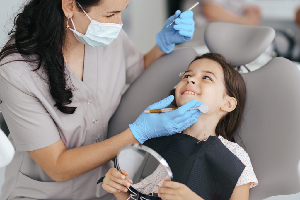 Mała dziewczynka siedzi na fotelu dentystycznym, trzymając w ręce lusterko